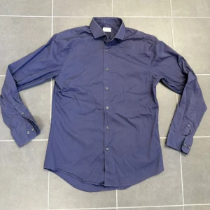 Blå skjorta från Tiger of Sweden - Snygg blå skjorta från Tiger of Sweden i slim fit-modell. Skjortan har klassisk krage, långa ärmar med manschett och knäppning framtill. Perfekt för dig som gillar stilrena och enkla plagg med en modern passform.