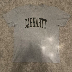 Grå Carhartt t-shirt XL - Snygg grå t-shirt från Carhartt i storlek XL med klassisk svart logotyp tryckt över bröstet. T-shirten har rund hals och korta ärmar, tillverkad i mjuk bomull som känns skön mot huden. Perfekt för dig som gillar streetwear och enkel stil.