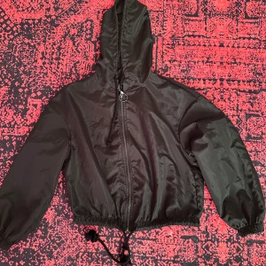 Svart vindjacka med huva och snörning - Svart vindjacka med huva, dragkedja med metallring och snörning i midjan. Jackan har elastiska muddar vid ärmslut och en loose passform. Perfekt för dig som gillar en enkel och stilren look.