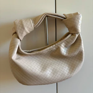 Beige axelväska i flätad look från Bubbleroom - Snygg beige axelväska från Bubbleroom med flätad struktur och coola knutdetaljer på axelbandet. Väskan har guldig dragkedja. Perfekt storlek för det viktigaste och ger en trendig touch till din outfit.