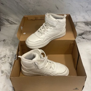 Vita höga sneakers med kardborre - Snygga vita sneakers med högt skaft och kardborreband upptill. Skorna har klassisk snörning och en platt sula. Perfekta för dig som gillar en clean och sportig look. 