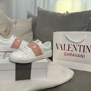 Valentino Rockstud sneakers vit/rosa - Snygga Valentino Rockstud sneakers i vitt skinn med breda ljusrosa band över vristen dekorerade med ikoniska nitar. Klassisk rund tå, vita snören och platt sula. Märkeslogga på plösen och insidan. Perfekta för dig som vill sticka ut med exklusiv streetstyle.