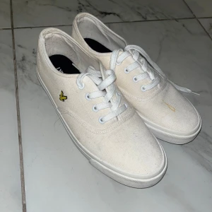 Vita sneakers från Lyle & Scott - Snygga vita sneakers från Lyle & Scott med klassisk låg profil och rund tå. Skorna är i canvas med vita snören och har den ikoniska gula fågeln broderad på sidan. Perfekta för en clean och stilren look till vardags.