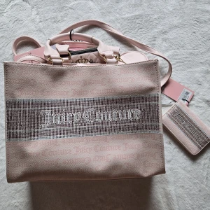 Rosa shoppingväska från Juicy Couture - Superfin rosa shoppingväska från Juicy Couture med glittriga detaljer och logga framtill. Väskan har både handtag och axelrem, samt matchande liten plånbok. Insidan är fodrad med tryckt logomönster och har dragkedjefack. Perfekt för dig som vill sticka ut med en trendig och rymlig väska.