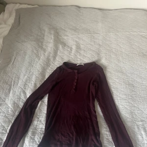 Vinröd långärmad tröja Pull&Bear - Säljer en vinröd långärmad tröja från Pull&Bear. Tröjan har rund halsringning och knappar framtill, vilket ger en enkel men snygg look. Materialet är ribbat och stretchigt, perfekt för lager-på-lager under hösten och vintern.