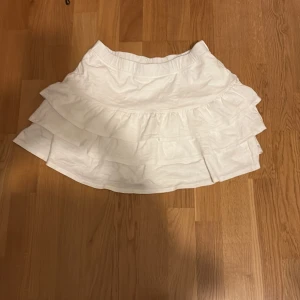 Vit volangkjol med lager - Supersöt vit kortkjol med flera volanger som ger en fluffig och lekfull look. Perfekt för dig som gillar en feminin och trendig stil. Kjolen har resår i midjan för extra komfort och är tillverkad i ett lätt tyg.