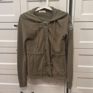 Olivgrön hoodie från Hollister XS - Olivgrön hoodie från Hollister med huva, snörning i midjan och dragkedja framtill. Tröjan har långa ärmar, ribbade muddar och en patch på ena ärmen. 