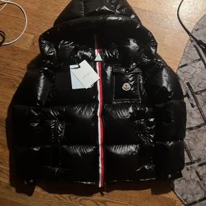 Moncler Montbeliard - Snygg svart pufferjacka från Moncler med glansig finish, huva och klassisk logga på bröstet. Jackan har rödvit rand längs dragkedjan, praktiska fickor och coolt innerfoder med serietidningsmotiv. Perfekt för kalla dagar när du vill hålla stilen. Använd December 2024 till januari 2025. Säljer den då det är dags att uppgradera till en ny jacka för vintern. Storlek 3/L