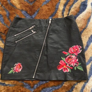 Svart skinnkjol med broderade blommor - Cool svart kortkjol i skinnimitation med broderade röda och rosa blommor framtill. Kjolen har snedställd dragkedja och två dekorativa dragkedjor upptill. Snygg passform och detaljer som ger en edgy vibe.