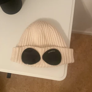 C.P. Company mössa äkta - Äkta nyskick knappt använd. Säljer en ikonisk vit ribbstickad mössa från C.P. Company med svarta goggle-linser framtill. Mössan är tillverkad i mjuk ull och har en bred uppvikt kant. Perfekt för dig som vill sticka ut med streetwear-vibe. Nypris 1500kr. Storlek M i ungsomstorlek men passar som strl. S i vuxen. Köpt i NK i sthlm