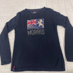Mörkblå stickad tröja från Morris - Mörkblå stickad tröja från Morris med broderad Union Jack-flagga och lejon på bröstet samt tydlig MORRIS-text. Rund halsringning och långa ärmar. Perfekt för dig som gillar klassisk brittisk stil och snygga detaljer.