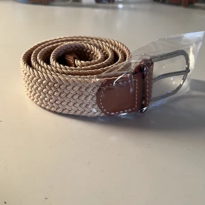  Beige flätat bälte  - Snyggt beige flätat bälte med bruna skinn-detaljer och silvrigt spänne. Bältet har en stilren look med tydlig struktur i materialet och passar perfekt till jeans eller chinos. Klassisk design som lyfter vilken outfit som helst. Bältet är elastiskt, detta bidrar till bättre passform. Det kan gå upp till 150cm.