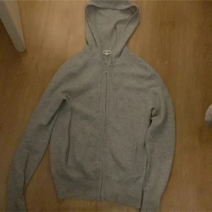 Grå hoodie i kashmir från Soft Goat XS - Grå hoodie från Soft Goat i mjuk kashmir. Tröjan har huva med snörning, dragkedja framtill och långa ärmar. Perfekt för chill dagar och snygg att ha under en väst. Enkel och stilren design med ribbade muddar. Den har 2 mini hål en finns på bild på armen och en vid rygg/axeln. 