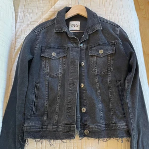Svart jeansjacka från Zara - Säljer en svart jeansjacka från Zara med snygga slitningar och rå kant nedtill. Jackan har klassisk krage, två bröstfickor med knappar och långa ärmar. Perfekt för dig som gillar en cool och avslappnad stil.