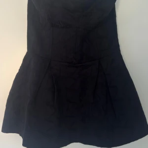 Svart axelbandslös top - Snygg svart axelbandslös top med utställd kjol och markerad midja. Tyget har ett subtilt mönster som ger extra attityd. Perfekt för dig som gillar en stilren look med lite edge. Topen  är kort och har en feminin siluett.