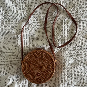 Handväska i flätad strå  - Söt rund axelremsväska/handväska i mörkt flätat strå. Mått: 16 cm i diameter, ca 6,5 cm djup 