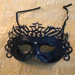 Svart glittrig maskeradmask - Snygg svart maskeradmask med glittriga detaljer och elegant utskuren design. Masken har svarta band för att knyta runt huvudet och dekorativa silvriga nitar framtill. Perfekt för maskerad eller temafest.