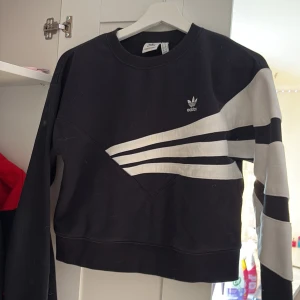 Svart och vit Adidas sweatshirt - Svart sweatshirt från Adidas med vita diagonala ränder och logga på bröstet. Tröjan har lång ärm med vita paneler och en croppad passform. Tillverkad i mjuk bomull, perfekt för en sportig och chill stil.