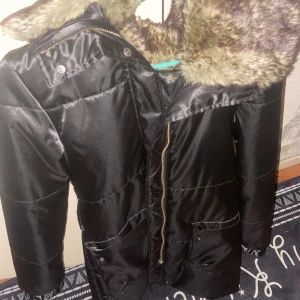 Svart pufferjacka med pälsluva - Säljer en svart pufferjacka med stor fluffig luva i brunmelerad fuskpäls. Jackan har dragkedja framtill, två stora fickor med knappar och quiltade detaljer. Perfekt för kalla dagar när du vill vara både varm och snygg.