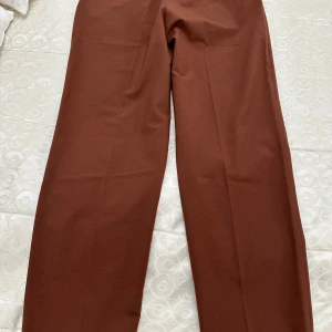 Bruna kostymbyxor med raka ben - Snygga bruna kostymbyxor med klassisk rak passform och pressveck. Byxorna har hög midja och stängs med knapp och dragkedja. Materialet känns mjukt och har en lätt glans.