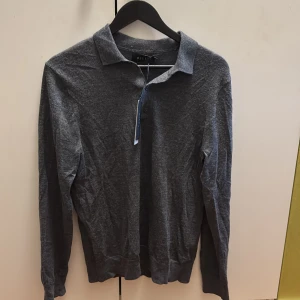 Mörkgrå pikétröja från Riley - Snygg mörkgrå pikétröja från Riley med klassisk krage och knäppning framtill. Långärmad modell i mjukt bomullsmaterial, perfekt för en stilren och avslappnad look.