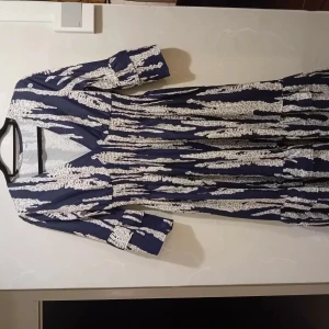 Marinblå långklänning med leopardmönster - Långklänning i marinblått med vita och gråa leopardmönstrade detaljer. Klänningen har v-ringning, trekvartsärm och smockad midja som ger en snygg siluett. Materialet känns lätt och flowy, perfekt för dig som gillar statement-plagg.