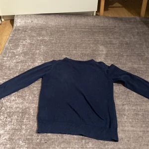 Mörkblå stickad tröja från GANT - Snygg mörkblå stickad tröja från GANT med rund halsringning och diskret logga på bröstet. Tröjan har långa ärmar och ribbade muddar vid ärmslut och nederkant. Perfekt för lager-på-lager under hösten och vintern.