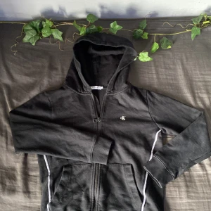 Svart hoodie från Calvin Klein Jeans - Svart hoodie från Calvin Klein Jeans med dragkedja framtill och liten CK-logga på bröstet. Tröjan har huva, långa ärmar och två fickor framtill. Tillverkad i mjuk bomull, perfekt för chill dagar eller när du vill ha en clean look.