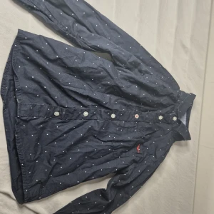 Mörkblå prickig skjorta Hampton Republic - Snygg mörkblå skjorta från Hampton Republic med vita prickar och klassisk krage. Skjortan har långa ärmar, knappar framtill och en liten röd broderad detalj på bröstfickan. Perfekt för dig som gillar stilrena men ändå lekfulla plagg.