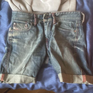Jeansshorts från Diesel, storlek 25 - Snygga jeansshorts från Diesel i klassisk blå denim med uppvikta benslut och röda sömmar. Shortsen har fem fickor, coola detaljer på bakfickorna och är tillverkade i Italien av bomull. Perfekta för en chill sommarlook. Kostar ca 1000-2000 som nya