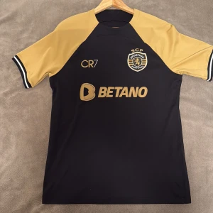 Ronaldo fotbollströja Nike  - Sporting CP fotbollströja i svart och guld med CR7-tryck och nummer 28 på ryggen. Tröjan har korta ärmar, broderat klubbmärke och är tillverkad i Dri-FIT-material som andas. Snygga detaljer med sponsorloggor och klassisk Nike-design. pris kan diskuteras