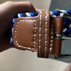 Randigt flätat bälte med skinn och blå detaljer - Snyggt flätat bälte med bruna skinnpartier och silverspänne. Bältet har ett randigt mönster i svart, blått, vitt och grått. Perfekt accessoar för att lyfta din outfit med lite färg och stil. Klassisk design med robust känsla och tydliga kontrastsömmar på skinnet.