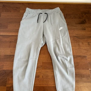 Grå/ljusblå mjukisbyxor från Nike - Snygga grå mjukisbyxor från Nike med svart snörning i midjan och diskret logga på vänster ben. Byxorna har elastisk midja, smal passform och praktiska fickor, varav en med dragkedja bak. Perfekta för chill eller träning.