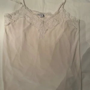 Beige spetslinne från Amisu - Säljer ett ljusbeige linne från Amisu med tunna axelband och vacker spetsdetalj längs urringningen och ryggen. Toppen har en feminin v-ringning och är tillverkad i ett mjukt, lätt material som känns skönt mot huden.
