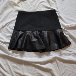 Svart volangkjol från Zara Woman - Supersnygg svart kortkjol från Zara Woman i storlek S. Kjolen har en volangkant i skinnimitation som ger en cool och trendig look. Hög midja och figurnära passform upptill, perfekt att styla med strumpbyxor och boots.