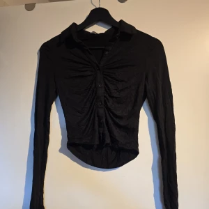 Svart croppad skjorta med knappar - Snygg svart croppad skjorta med lång ärm och krage. Skjortan har knappar framtill och ett rynkat parti som ger en cool och trendig look. Perfekt för dig som gillar att sticka ut med stilrena detaljer och figurnära passform. Använd 1 gång