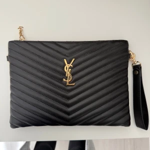 Svart clutch väska från YSL - Snygg svart clutch väska från YSL med guldiga detaljer och logotyp framtill. Väskan är quiltad i ett chevronmönster och har en praktisk handledsrem samt dragkedja med YSL-hänge. Perfekt för dig som vill ha en stilren och lyxig accessoar.