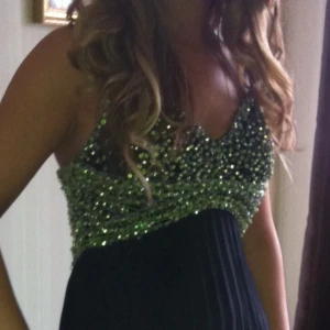 Glittrig långklänning med paljetter - Snygg långklänning med glittrig överdel täckt av gröna paljetter och djup v-ringning. Kjolen är svart, lång och plisserad vilket ger ett elegant fall. Perfekt för dig som vill sticka ut med en glamorös look.