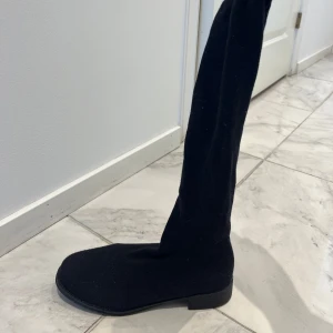 Svarta stretchiga stövlar - Svarta, höga stövlar med platt sula och rund tå. Skorna är gjorda i ett stretchigt tygmaterial som sitter tight längs benet. Perfekta för en stilren look och enkla att matcha med olika outfits.