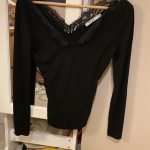 Svart ribbad topp med spets från NA-KD - Säljer en svart långärmad topp från NA-KD med ribbad struktur och snygg v-ringning dekorerad med spetsdetaljer. Toppen har en figurnära passform och är perfekt för dig som gillar stilrena plagg med en twist.