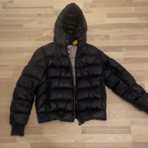 Svart dunjacka Parajumpers Sheen M - Säljer en svart dunjacka från Parajumpers, modell Sheen. Jackan har en puffig look, huva, dragkedja och coolt PJS-märke på ärmen. Insidan är ljusgrå och jackan har ribbade muddar. Perfekt för kalla dagar och riktigt snygg streetstil.