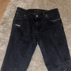 Svarta jeans från Nudie jeans - Svarta jeans från Nudie Jeans i slim fit med slitningar på ena låret och vid fickan. Klassisk femficksmodell med dragkedja och knapp. Jeansen har mörk tvätt och är tillverkade i stretchig bomullsdenim för en tajt passform.