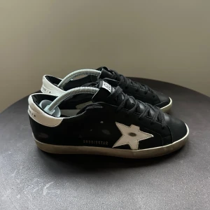Golden Goose svarta sneakers med stjärna - Svarta sneakers från Golden Goose med vit stjärndetalj på sidan och vit hälkappa. Skorna har snörning, platt sula och klassisk rund tå. Snygg kontrast mellan svart och vitt, samt diskreta logodetaljer på sidan. Perfekta för en cool och avslappnad stil.