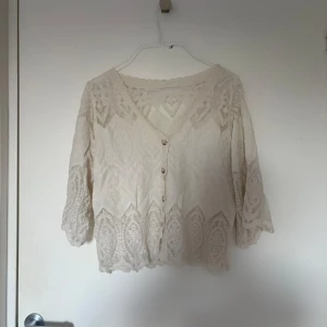 Beige spetsblus med knappar - Säljer en romantisk beige blus i spets med v-ringning och trekvartsärmar. Blusen har fina broderade detaljer, knappar framtill och en lätt croppad passform. Perfekt för dig som gillar boho-stil och vill ha något luftigt och snyggt.