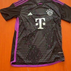Svart FC Bayern München Adidas tröja - Säljer en svart FC Bayern München fotbollströja från Adidas med lila och gröna diamantmönster på framsidan. Tröjan har korta ärmar, lila detaljer längs sidor och axlar samt broderat klubbmärke och Adidas-logga. Materialet är lätt polyester med Aeroready-teknologi.