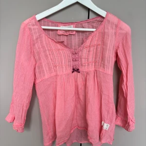 Rosa blus från Odd Molly med spets - Supersöt rosa blus från Odd Molly med tunna ränder, virkade spetsdetaljer och dekorativa knappar framtill. Blusen har v-ringning, trekvartsärm och en liten rosett. Luftigt material som passar perfekt till vår och sommar.