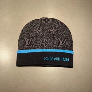 Svart LV bälte - Snygg svart mössa från Louis Vuitton med klassiskt monogrammönster i grått och en blå kant längst ner. Märkets logga är broderad i blått framtill. Perfekt accessoar för dig som vill sticka ut med lyxig streetstyle. Materialet är stickad och mjukt.