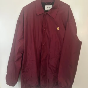 Vinröd fodrad vindjacka från Carhartt L - Säljer en vinröd vindjacka från Carhartt Work In Progress i storlek L. Jackan har klassisk krage, tryckknappar framtill, två sidofickor och Carhartt-logga i gult på bröstet. Perfekt för dig som gillar streetwear och enkel stil.