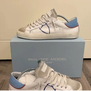 Philippe Model vita sneakers med blå detalj - Snygga vita Philippe Model sneakers med blå och röda detaljer vid hälen och beige mocka framtill. Skorna har klassisk snörning, platt sula och en blå sköldlogga på sidan. Perfekta för dig som gillar stilrena men unika sneakers. Original snöre finns ej inga sulor box ingår ej 