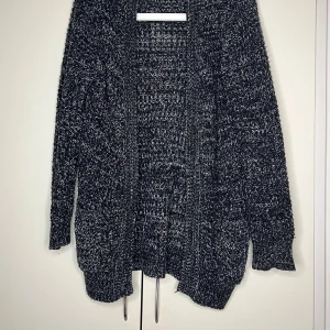 Svartvit stickad kofta - Mysig svartvit stickad kofta med grov struktur och öppen front. Koftan har lång ärm och en avslappnad passform, perfekt att slänga på sig när det är kyligt. Den melerade looken ger en cool och avslappnad vibe.